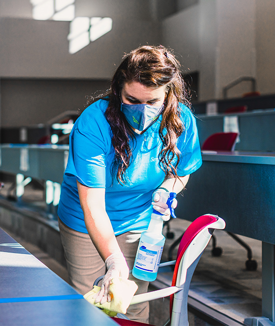 Two cleaning people are cleaning the inside of a classroom by wiping down desks and using a microfiber mop to wipe dust and debris off floors
