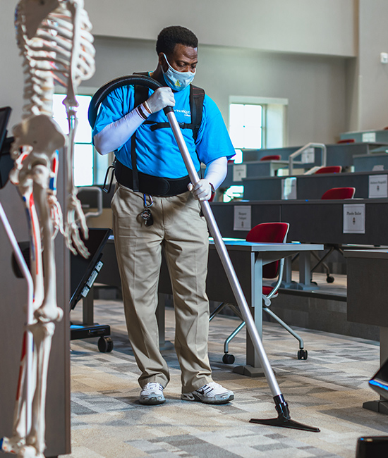 Two cleaning people are cleaning the inside of a classroom by wiping down desks and using a microfiber mop to wipe dust and debris off floors